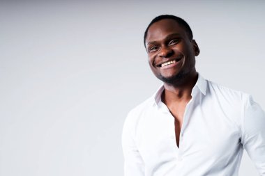 Beyaz gömlekli, açık yakalı, mutlu, esmer adam kameraya bakıp gülümsüyor.