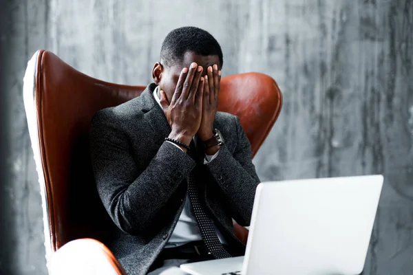 Unavený africký americký kancelářský pracovník sedí na židli u notebooku a zakrývá si obličej rukama — Stock fotografie