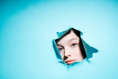 Güzel makyajlı bir kız parlak bir arka plandaki delikten kameraya bakar.