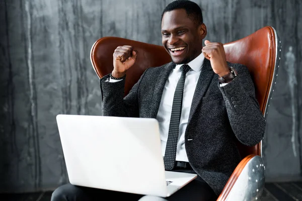 Afrykański amerykański biznesmen siedzi na krześle i pracuje na laptopie radując się umową — Zdjęcie stockowe