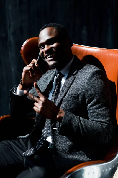 Bonito empresário afro-americano sentado na cadeira e falando ao telefone mostrando sinal de paz — Fotografia de Stock