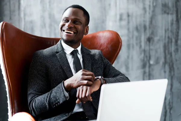 Feliz empresário afro-americano de terno sentado em uma cadeira e trabalhando em um laptop olhando para o relógio em sua mão — Fotografia de Stock