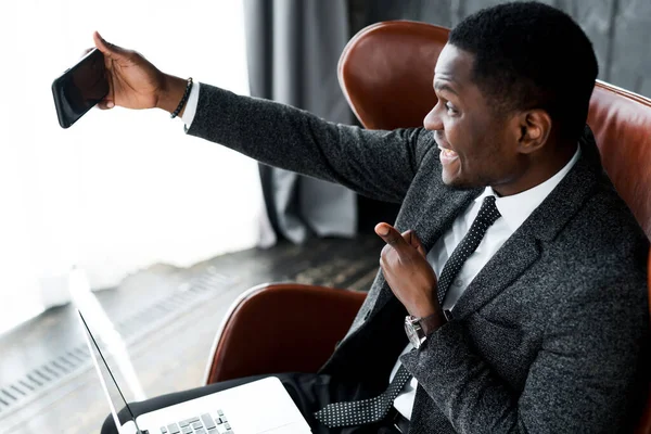 Glimlachende Afrikaanse Amerikaanse zakenman zitten op stoel met laptop en het nemen van selfie tonen duim omhoog — Stockfoto