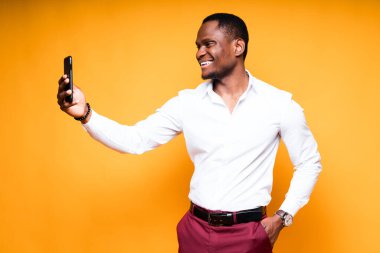 Tişörtlü, mutlu, esmer bir adam elinde telefonla selfie çekip gülümsüyor.