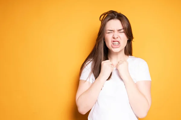 Een meisje met een beugel, die haar ogen dicht doet van de pijn, haar handen in vuisten klemt en doet alsof ze zichzelf in de kaak slaat. — Stockfoto