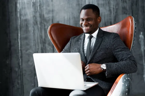 Улыбающийся африканский американский бизнесмен сидит на стуле и проводит деловую конференцию на ноутбуке — стоковое фото