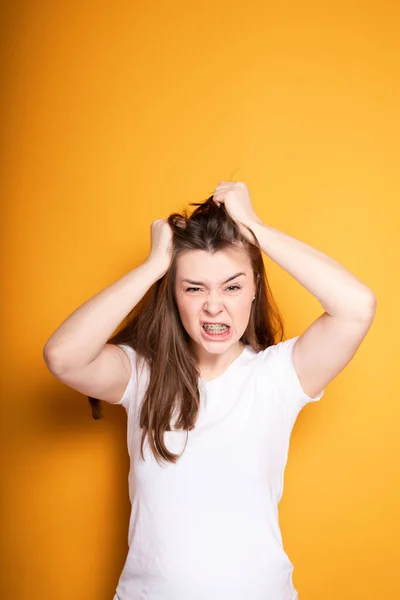 Wściekła młoda dziewczyna z aparatem na zębach wyciąga włosy z głowy od bólu zębów i głowy stojąc na żółtym tle — Zdjęcie stockowe