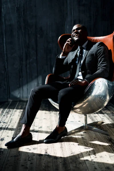 Elegante empresário afro-americano sentado na cadeira e falando ao telefone sorrindo — Fotografia de Stock