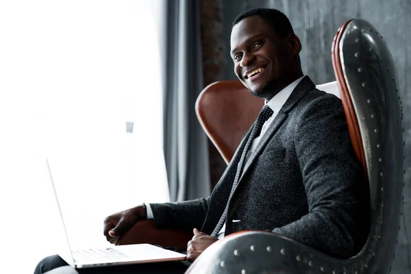 Ευτυχισμένος όμορφος Αφροαμερικάνος υπάλληλος γραφείου που κάθεται στο γραφείο και χαμογελά ενώ εργάζεται στο laptop — Φωτογραφία Αρχείου