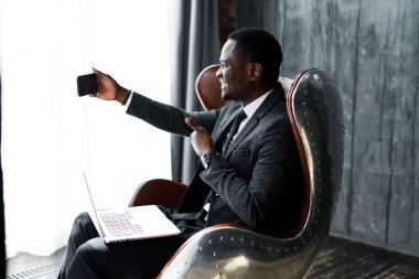 Yakışıklı afro-amerikan iş adamı dizüstü bilgisayarda çalışıyor ve telefonda selfie çekiyor.