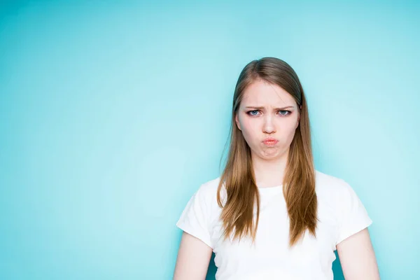 Ένα χαριτωμένο λυπημένο κορίτσι με λευκό μπλουζάκι βγάζει τα χείλη της από πικρία. Απομονωμένα σε μπλε φόντο — Φωτογραφία Αρχείου