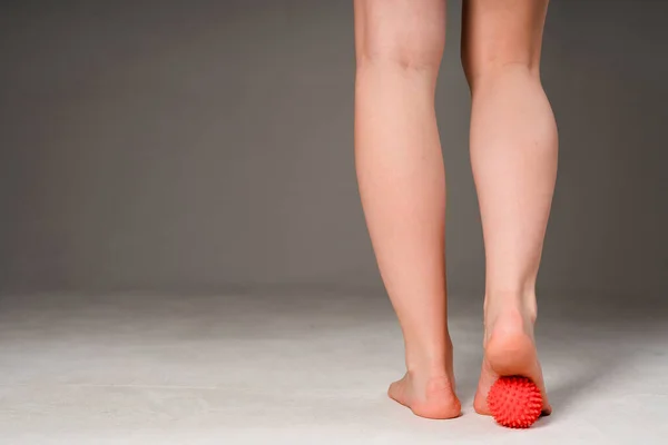 Photo of female sports legs. A woman does a foot massage with a red massage ball. Isolated on gray background