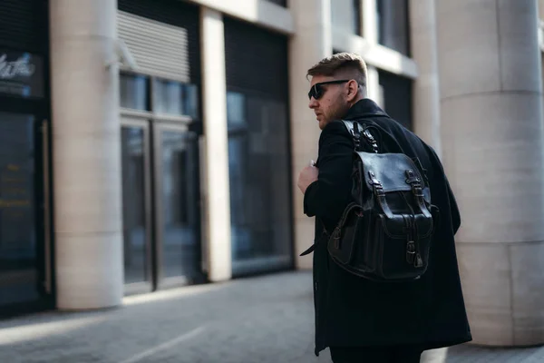 Красивый турист в солнечных очках и черном пальто с рюкзаком на одном плече ходит по городу и смотрит в сторону. Фото сзади — стоковое фото