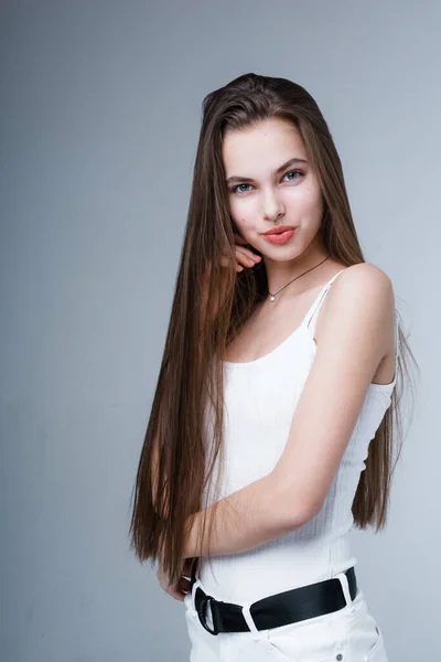 Rapariga atraente em um top de tanque branco e calças com cabelo longo e brilhante fica de lado e olha para a câmera — Fotografia de Stock