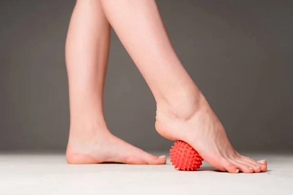 Photo of female sports legs. A woman does a foot massage with a red massage ball. Isolated on gray background