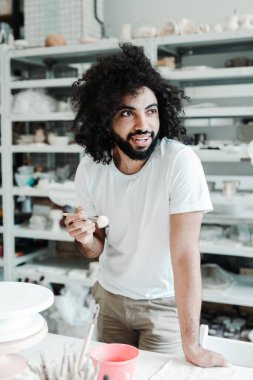 Kıvırcık saçlı Türk, yaratıcı bir stüdyoda elinde boya fırçasıyla duruyor. Gülümsüyor ve başka tarafa bakıyor.