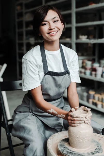 Una mujer asiática linda se sienta a la rueda de un alfarero en un estudio de hobby y hace un jarrón de barro. Hermosa sonrisa, dientes blancos — Foto de Stock