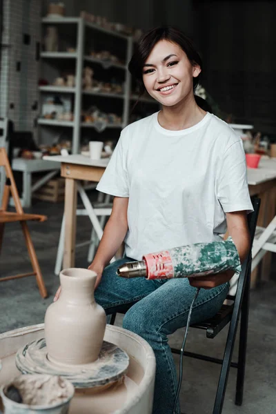 Een Aziatische vrouw droogt een kleivaas met een bouwhaardroger in een pottenbakkerij. houdt zich bezig met een hobby — Stockfoto