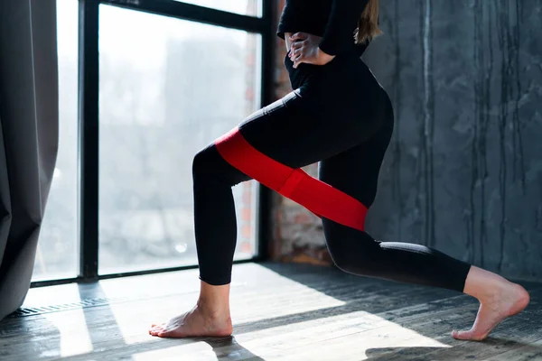 Flickan är engagerad i gymmet och gör knäböj på ett ben med hjälp av ett elastiskt band. Övningar med sportutrustning — Stockfoto