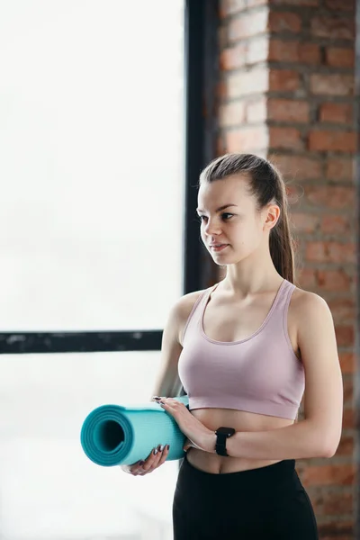 En vacker flicka tränare i en sport topp och leggings håller en yogamatta i händerna när hon står mot bakgrund av fönster — Stockfoto