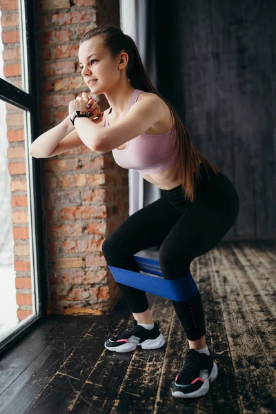 Ung charmig flicka i gymmet gör knäböj med hjälp av ett elastiskt band. Friska livsstil koncept — Stockfoto