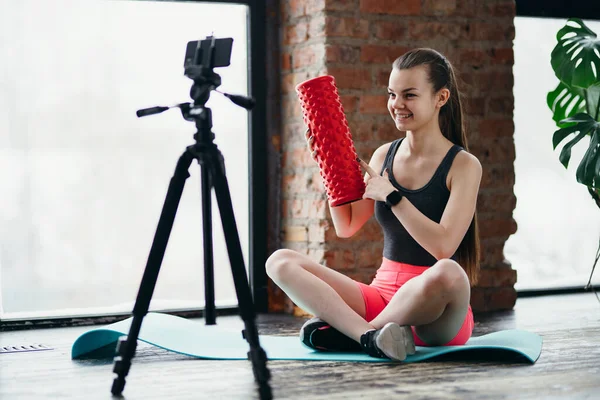 Дівчина фітнес-тренер сидить на килимку для йоги і тримає фасцію в руках під час запису відео на камеру на тринозі — стокове фото