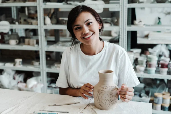 Une jolie fille asiatique dans un T-shirt blanc s'assoit à une table et sourit magnifiquement dans un atelier de poterie et tient un vase en argile fait maison dans sa main. Hobby concept de studio. — Photo