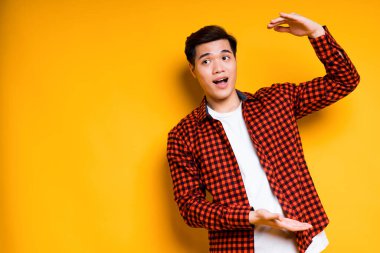 Tişörtlü ve tişörtlü mutlu Asyalı adam elleriyle büyük bedenini gösteriyor ve şaşırıyor. Sarı arkaplanda izole edilmiş