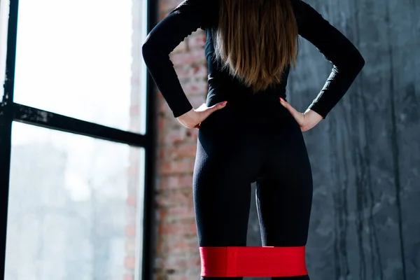 Photo from behind. The girl rests her hands on her hips and does exercises on her legs using an elastic band — Stock Photo, Image