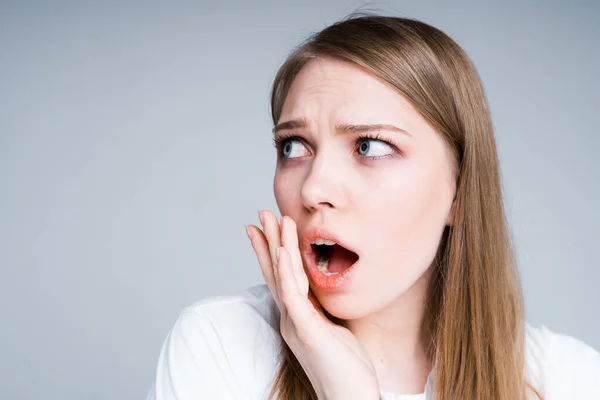 Une jolie fille dans un T-shirt blanc regarde sur le côté, surprise et indignée, ouvrant sa bouche et mettant sa main à sa bouche — Photo