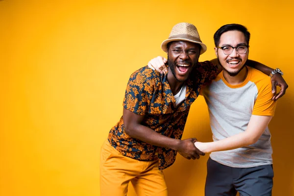 Cheerful African American guy in a hat and shirt hugs a happy Asian guy. Meeting friends after a long separation