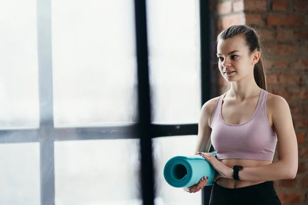 Flicka fitness tränare i en sport topp och leggings förbereder sig för ett träningspass hålla en yogamatta i händerna — Stockfoto