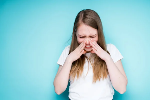 Una ragazza carina con una maglietta bianca è malata di raffreddore e starnutisce, chiudendo gli occhi e toccandosi il naso con le mani — Foto Stock