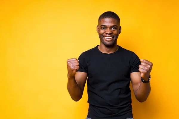 Szczęśliwy Afroamerykanin w czarnej koszulce uśmiecha się szeroko i raduje się zwycięstwem zaciskając dłonie w pięści — Zdjęcie stockowe