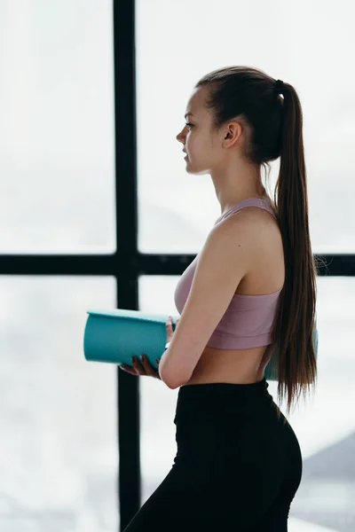 Muchacha atractiva en ropa deportiva sostiene una esterilla de yoga en sus manos y se prepara para un entrenamiento mientras está de pie de lado —  Fotos de Stock