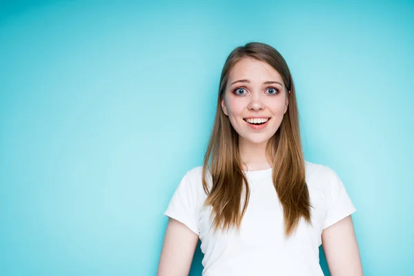 Ένα χαρούμενο κορίτσι με λευκό μπλουζάκι είναι έκπληκτο και χαρούμενο ενώ στέκεται σε μπλε φόντο και ανοίγει το στόμα της — Φωτογραφία Αρχείου