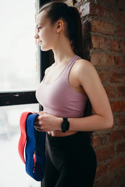 Una hermosa chica linda en una parte superior sostiene bandas elásticas para el entrenamiento en sus manos y mira por la ventana — Foto de Stock