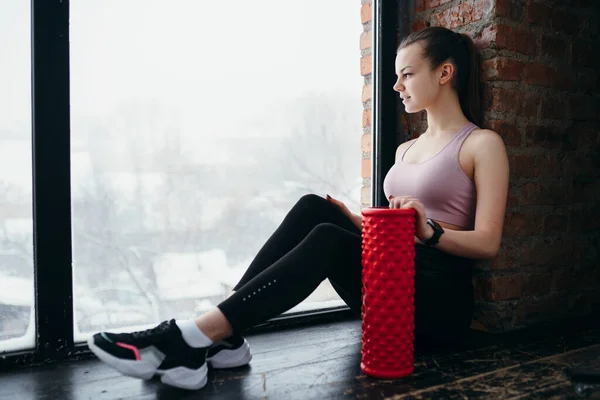 Chica entrenador sentado en el suelo en el gimnasio junto a la fascia en el fondo de las ventanas preparándose para un entrenamiento —  Fotos de Stock