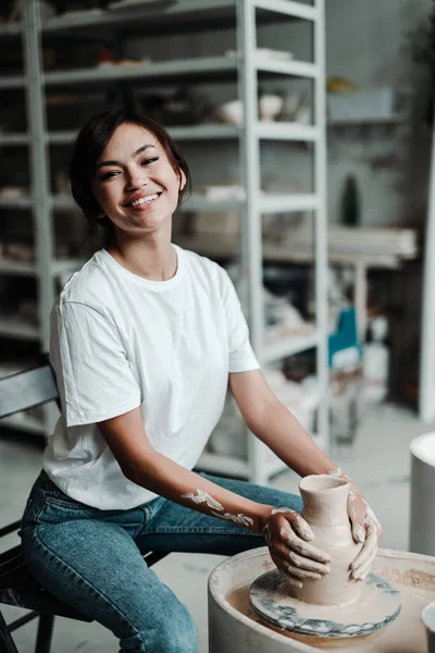 Heureuse fille asiatique dans un t-shirt et un jean assis sur une chaise fait des plats en argile avec ses mains — Photo