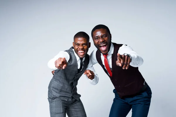 Dwóch wesołych afrykańskich biznesmenów w koszulkach i kamizelkach śmieje się i wytyka palcami kamerę. — Zdjęcie stockowe