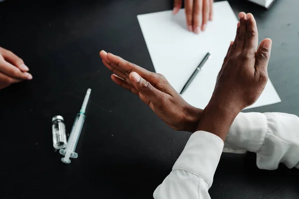 Photo from above. Hands of an African American woman protesting and refusing the vaccine lying on the table, crossing her palms