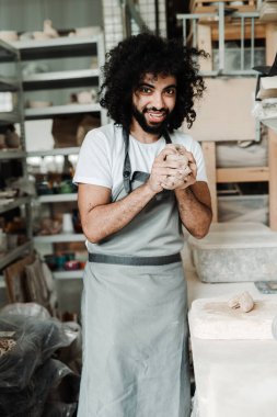  Kıvırcık saçlı bir adam, elleri bir hobi stüdyosunda önlük ve beyaz bir tişörtle çömlek kilini buruşturur.