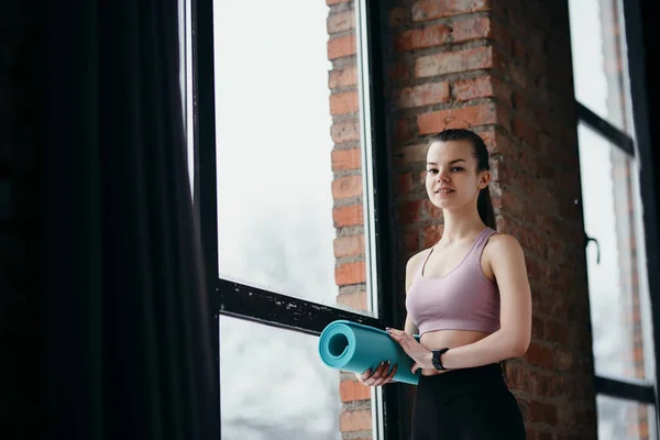 Мила молода дівчина фітнес-тренер тримає йогу килимок в руці, стоячи в спортзалі і готуючись до тренувань — стокове фото