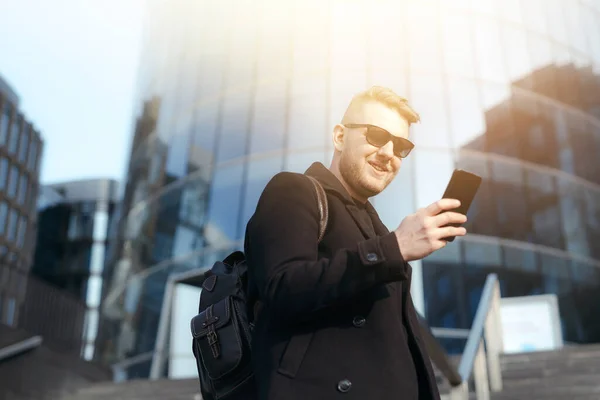 Мужчина в бороде и солнцезащитных очках держит в руках телефон и пишет сообщение возле бизнес-центра — стоковое фото