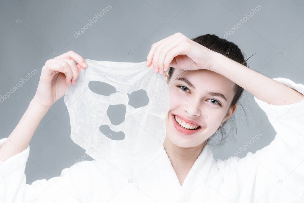  A cute girl in a white velvet robe holds a cosmetic mask and smiles on a gray background. cosmetology concept