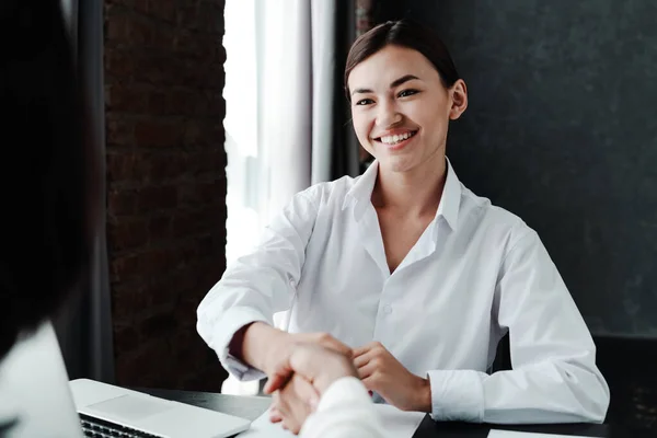 Молодая улыбающаяся азиатская девушка, сидящая за столом, заканчивает переговоры пожимая руку женщине, которая берет ее на работу — стоковое фото