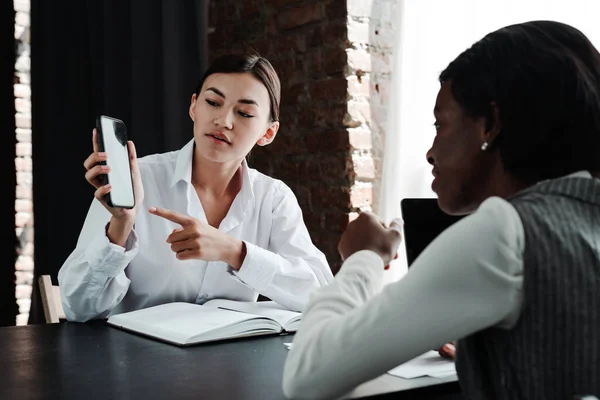 Young asian girl holding a phone in her hand and advertises it to african american woman sitting at the table