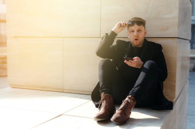 Yakışıklı bir adam binanın önünde oturuyor ve elinde güneş gözlüğü ile elinde bir telefon tutuyor.