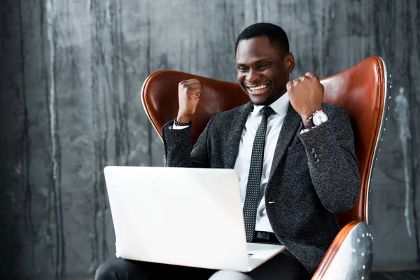 Африканский американский бизнесмен, сидящий на стуле и работающий за ноутбуком, радуясь сделке — стоковое фото