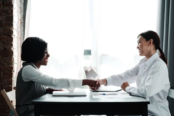 Afrykańska amerykańska bizneswoman i azjatka siedzą przy stole naprzeciwko siebie, uściskując sobie dłonie, uśmiechając się — Zdjęcie stockowe
